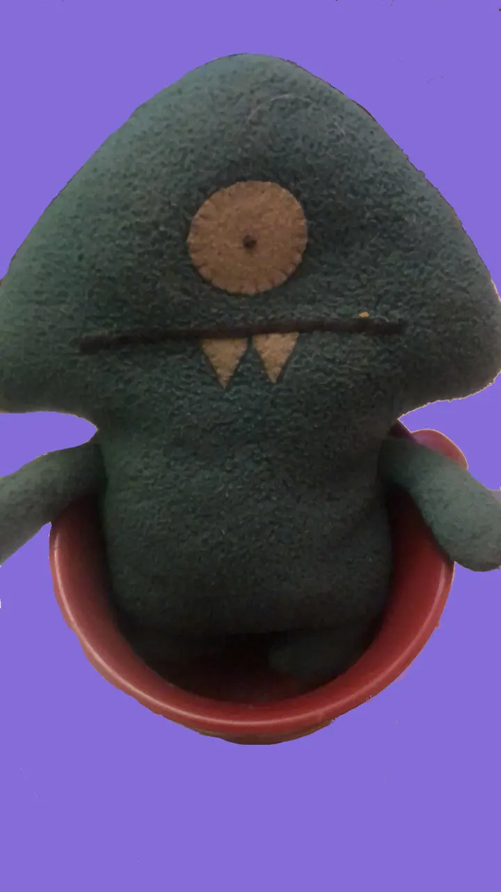 A green felt puppet with one eye and two fangs sits in a red ceramic mug against a lavender background