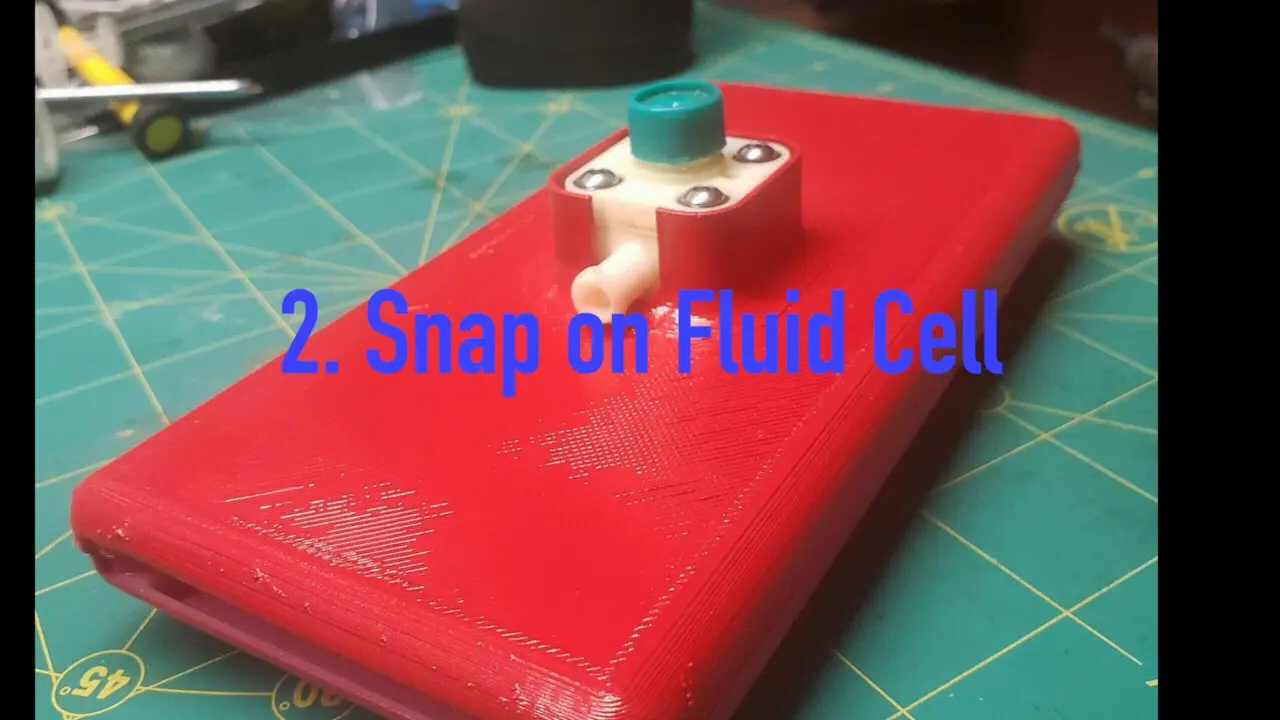 A red 3D-printed handheld with a white fluid cell and teal cap on a green cutting mat, captioned 2. Snap on Fluid Cell