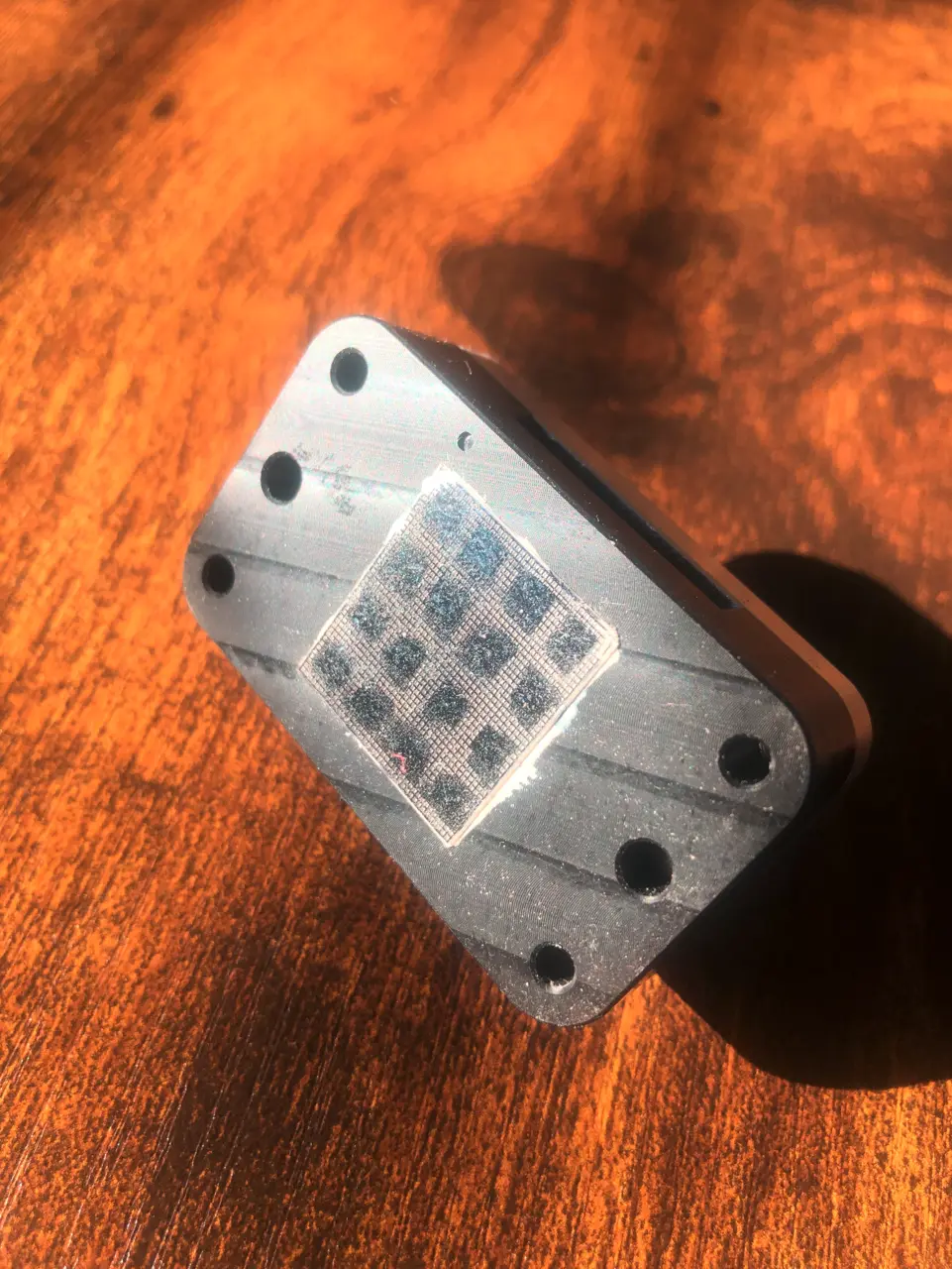 A square silicon chip with a four by four grid of windows held in a machined aluminium carrier with four screw holes