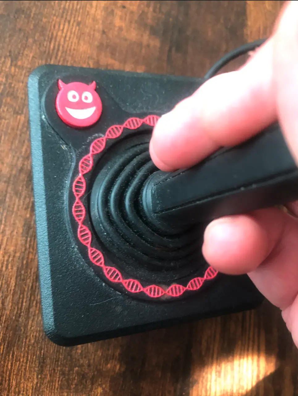 A hand on a black joystick with a red cartoon demon badge and a red DNA helix printed around the stick